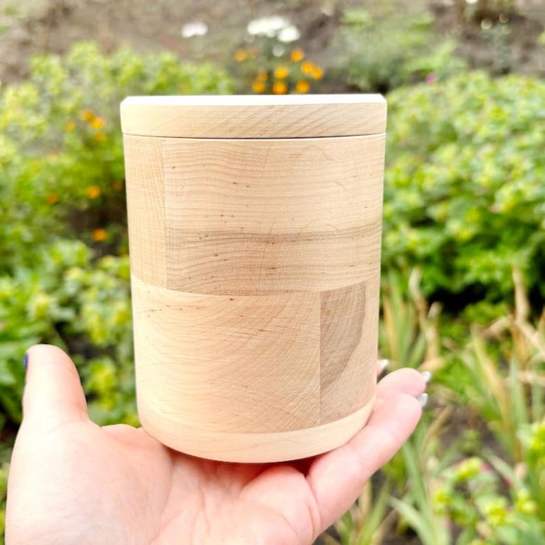 K&ouml;nnte beinhalten: Ein zylindrischer Holzbeh&auml;lter mit Deckel, gehalten in einer Hand. Der Beh&auml;lter besteht aus hellem Holz mit sichtbarer Maserung und einem geometrischen Muster. Der Deckel sitzt b&uuml;ndig oben. Der Hintergrund ist mit gr&uuml;nem Laub verschwommen.