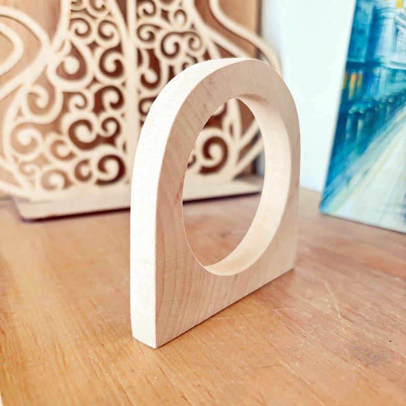 May include: A light-colored, smooth wooden arch with a circular cutout in the center. The object has a flat base and a curved top, featuring a natural wood grain pattern throughout its surface. The structure is solid and stands upright on its own.