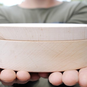 Può includere: Una scatola rotonda di legno chiaro con coperchio. La scatola è tenuta tra le mani di qualcuno e la venatura del legno è visibile. La scatola sembra nuova e inutilizzata. La scatola ha un diametro di circa 13 cm.