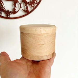 Può includere: Una piccola scatola rotonda di legno chiaro con coperchio, tenuta in mano. Il legno presenta venature naturali. Il coperchio si adatta perfettamente. La scatola misura circa 7,6 cm di diametro e 7,6 cm di altezza.