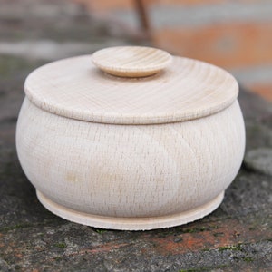 May include: A small, round, wooden box with a lid. The box is made of light-colored wood and has a smooth finish. The lid is slightly raised and has a small knob on top.