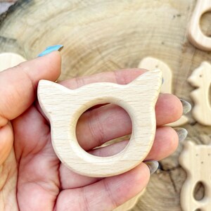 Peut inclure: Une bague de dentition en bois clair en forme de chat avec un centre circulaire. La bague a des bords arrondis et est tenue dans une main. D'autres formes en bois sont visibles en arri&egrave;re-plan.