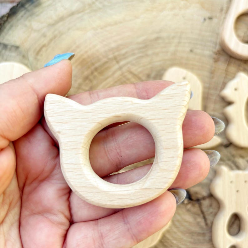 Peut inclure: Une bague de dentition en bois clair en forme de chat avec un centre circulaire. La bague a des bords arrondis et est tenue dans une main. D'autres formes en bois sont visibles en arri&egrave;re-plan.