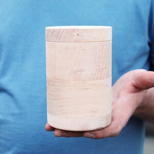 K&ouml;nnte beinhalten: Ein zylindrischer Holzbeh&auml;lter mit Deckel, der in der Hand einer Person gehalten wird. Der Beh&auml;lter besteht aus hellem Holz mit sichtbaren Maserungsmustern. Der Deckel passt fest auf. Der Hintergrund ist einfarbig blau.