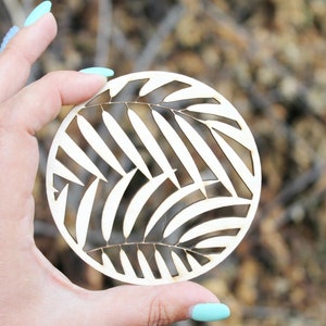 May include: A round wooden coaster with a laser-cut design of palm leaves. The coaster is white and the leaves are cut out, creating a negative space design.