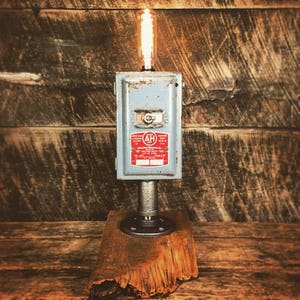 May include: A vintage industrial style lamp made from a repurposed electrical box with a red label that reads "Automatic Electric Heater Co.". The lamp is mounted on a black pipe and a wooden base.