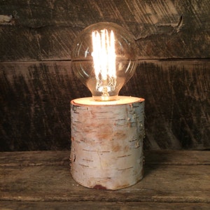 Può includere: Una base per lampada in legno di betulla con una lampadina in vetro trasparente. La lampadina è accesa e proietta un bagliore caldo.