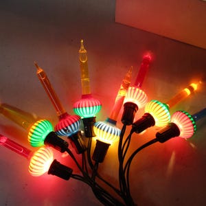 Może przedstawiać: Grupa zabytkowych lampek choinkowych z kolorowymi żarówkami. Żarówki mają unikalny design z żebrowaną, kulistą podstawą i długą, spiczastą końcówką. Światła są w różnych kolorach, w tym czerwonym, zielonym, żółtym i niebieskim.