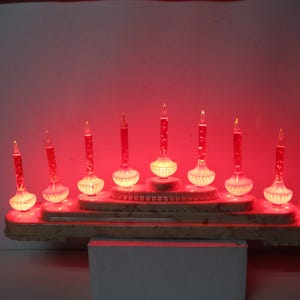 以下が含まれることがあります： 9つのキャンドル型のライトが点灯するヴィンテージのクリスマスデコレーション。 赤と白の電球を使用し、大理石模様の階段状のベースに配置されています。 ベースは長方形で、レトロな雰囲気です。