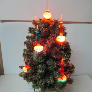 May include: A small artificial Christmas tree with metallic green needles, featuring several illuminated bubble lights in orange, blue, and red. The lights have ribbed bases and clear glass tubes, and the tree is set in a light-colored circular base.