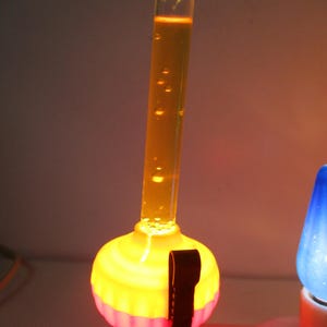 May include: A bubble light with a clear glass tube filled with orange liquid and small rising bubbles. The base is a bright yellow and pink fluted plastic housing with a black clip attached to the side, illuminated from within by a light source.