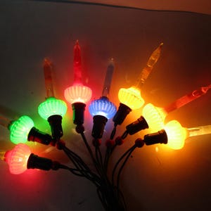 Może przedstawiać: Szereg kolorowych lampek choinkowych. Każda lampka ma żebrowaną, kulistą podstawę i przezroczystą, spiczastą końcówkę. Światła są czerwone, zielone, niebieskie i żółte. Światła są połączone czarnymi przewodami.