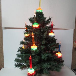 Może przedstawiać: Mała sztuczna choinka z kolorowymi światłami w kształcie świec. Drzewko ma zielone igły i jest ozdobione czerwonymi, pomarańczowymi, żółtymi i niebieskimi światłami. Drzewko stoi w beżowej doniczce.