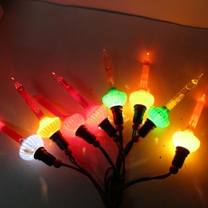 Może przedstawiać: Grupa zabytkowych lampek choinkowych z kolorowymi żarówkami. Żarówki są w różnych kolorach, w tym czerwonym, zielonym, żółtym i białym. Każda żarówka ma przezroczystą, spiczastą końcówkę. Lampki są na ciemnozielonym drucie.