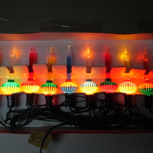 Pode incluir: Um fio de dez luzes de Natal de bolha em estilo vintage com bases de vidro caneladas e multicoloridas em tons de vermelho, verde, azul e amarelo, conectadas por um cabo elétrico preto e dispostas dentro de uma caixa de papelão.