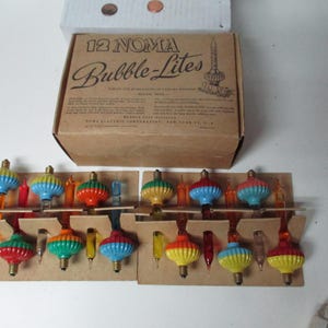 Może przedstawiać: Zestaw światełek choinkowych NOMA Bubble-Lites w stylu vintage. Pudełko jest brązowe z napisem "12 NOMA Bubble-Lites". Lampki są wielokolorowe, w tym czerwone, niebieskie, zielone i żółte. Lampki są na kartonowych uchwytach.