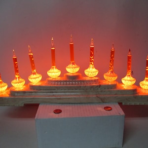 以下が含まれることがあります： 9つのオレンジ色のバブルライト電球が点灯する電気燭台。ベースは段状のマーブル模様のベージュのプラスチック製で、右側に電源コードが付いています。各電球は装飾的なガラスホルダーに収められています。