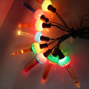 Może przedstawiać: Szereg zabytkowych lampek choinkowych z kolorowymi żarówkami. Żarówki mają żebrowany wzór i świecą w różnych kolorach, w tym czerwonym, zielonym, żółtym i niebieskim. Lampki połączone są czarnym przewodem.