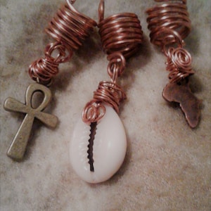 May include: Three copper wire hair beads with charms. The beads are coiled and have a decorative swirl at the top. The charms are an ankh, a cowrie shell, and a copper outline of the continent of Africa.