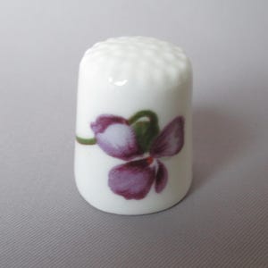 May include: A white porcelain thimble with a purple and green floral design. The thimble has a dimpled top and a cylindrical shape. The floral design features a purple flower with green leaves and stem. The thimble is a sewing accessory.