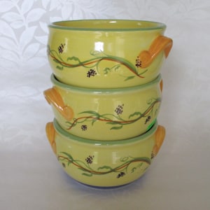 May include: Three yellow ceramic soup bowls with green and brown vine designs and yellow handles. The bowls are stacked on top of each other.