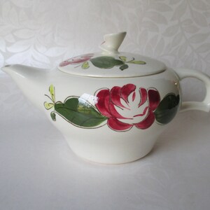 May include: White ceramic teapot with a floral design. The teapot has a lid with a knob and a handle. The floral design features red roses, green leaves, and yellow accents.