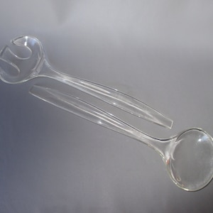 May include: Clear plastic salad serving set. The set includes a fork and a spoon, both with long, curved handles. The fork has three tines, and the spoon has a rounded bowl. The set is designed for serving salads.