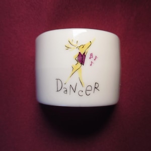 May include: White ceramic napkin ring with a whimsical illustration of a yellow deer in a dancing pose, wearing a purple vest. The word "Dancer" is written below the deer in black script. Musical notes are also present.
