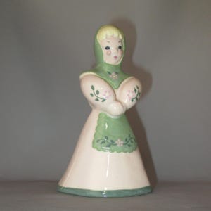 May include: A ceramic figurine of a girl wearing a green dress with pink flowers and a green hood. The figurine has a white face with pink cheeks and black eyes.