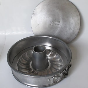 May include: A vintage, round, aluminum Bundt cake pan with a removable lid. The pan features a central tube and a fluted design. The lid has the text "Dr. Oetker Made in Germany".
