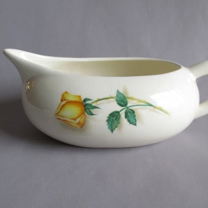 May include: A white ceramic gravy boat with a yellow rose design. The rose has green leaves and a stem with thorns.