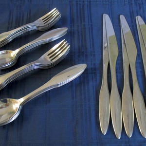 May include: A collection of stainless steel silverware, including forks, spoons, and knives, arranged on a dark blue surface. The silverware has a simple, elegant design, reflecting light. The image showcases a complete set of dining utensils.