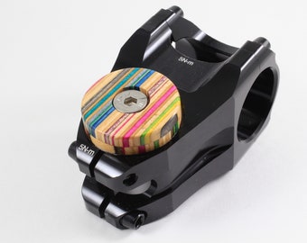 May include: Black bicycle stem with a multi-coloured skateboard top cap. The stem has "5N-m" printed on the side.
