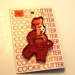 May include: A red translucent plastic chef-shaped cookie cutter attached to a white rectangular cardboard backing. The backing features the repeated red text "COOKIE CUTTER" and includes two small orange and white price stickers at the top.