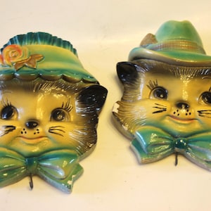 May include: Two vintage ceramic cat head wall hangings. One cat wears a green hat with a bow, the other a green bonnet with a flower. Both have yellow and brown fur, black eyes, and green bow ties.