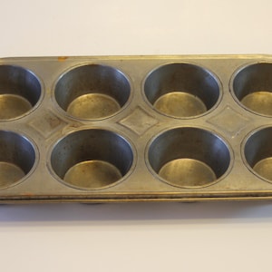 May include: An antique, rectangular muffin tin with eight circular molds. The metal tin has a worn, aged appearance with a light brown patina. The molds are arranged in two rows of four, with diamond-shaped accents between the rows.