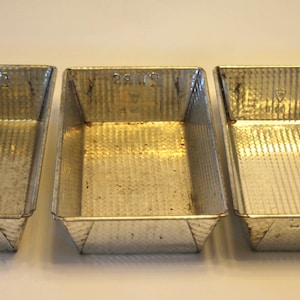 May include: Three vintage rectangular metal baking pans with textured sides and angled corners. The pans are made of a shiny, silver-colored metal and are empty. The pans are arranged side by side on a white surface.