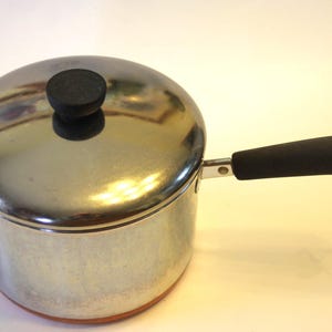 May include: A stainless steel saucepan with a lid and a black handle. The pot has a copper-colored bottom and a black knob on the lid. The handle has a metal loop at the end.