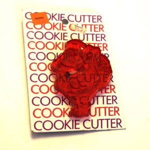 May include: A red plastic cookie cutter shaped like a Santa Claus figure, mounted on a white rectangular cardboard backing printed with the repeating text COOKIE CUTTER in red and purple, featuring two small orange and white price stickers.