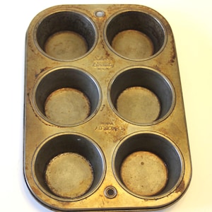 May include: A vintage six-cup muffin tin with a well-worn, aged appearance. The metal has a golden-brown patina with signs of rust. The words "Silver Beauty U.S.A." and "EKCOLOY" are stamped on the tin. A small hole is at the top and bottom.