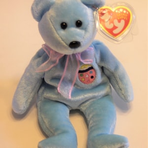 May include: A light blue beanie baby bear with a pink ribbon and an embroidered Easter egg on its chest. The bear has black eyes and a black nose. The bear is wearing a Ty Beanie Babies tag.