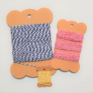 May include: Three spools of baker's twine wrapped around scalloped-edge cardboard cards. One large spool features blue and white twisted twine, a medium spool has pink and white twine, and a small spool contains yellow and white twine.
