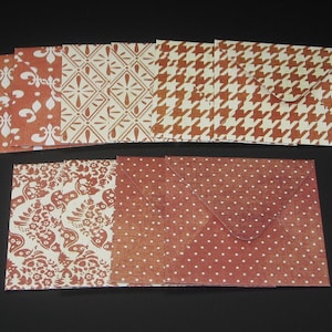 May include: A collection of square paper envelopes featuring various patterns in shades of terracotta and white. The designs include fleur-de-lis, geometric diamond motifs, houndstooth, intricate floral patterns, and small white polka dots on a solid background.