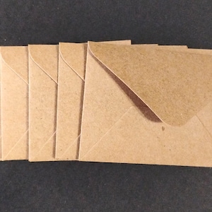 May include: A set of six rectangular brown kraft paper envelopes arranged in a slightly overlapping horizontal row. Each envelope features a pointed triangular flap on the top edge and a smooth, matte texture consistent with standard recycled paper material.