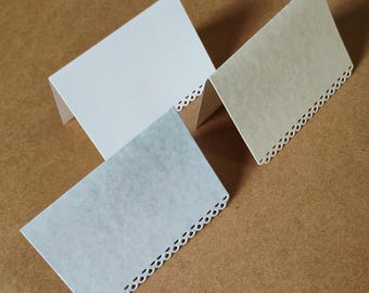 May include: Three folded paper place cards in white, cream, and light blue. Each card features a rectangular shape with a decorative die-cut scalloped edge along the bottom. The cards are positioned at different angles to show their folded structure.