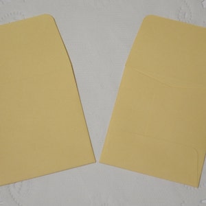 May include: Two rectangular envelopes in a pale yellow color. One envelope is positioned with the flap open and facing upward, while the other envelope is positioned with the flap folded down, revealing the back construction of the paper stationery.