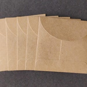 May include: A set of five square envelopes made from light brown kraft paper. Each envelope features a rounded flap closure and a smooth, matte texture. The envelopes are arranged in a slightly overlapping row, showcasing their uniform size and color.