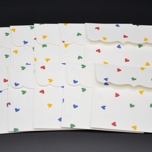 May include: A collection of ten white paper envelopes featuring a scalloped flap edge and a repeating pattern of small, scattered hearts in primary colors including red, blue, green, and yellow, arranged in two rows against a dark background.