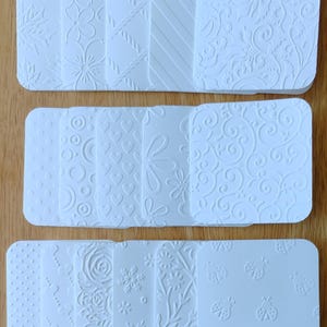 May include: A collection of white rectangular cards featuring various embossed patterns including floral designs, geometric shapes, diagonal lines, hearts, circles, snowflakes, and ladybugs. Each card has rounded corners and a textured surface.