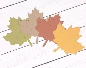 May include: Four paper leaves in different shades of green, brown, and yellow. The leaves are arranged in a cluster on a white wooden background.